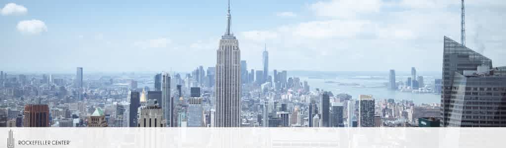 Panoramic view of New York City skyline from a high vantage point during daylight. Prominent skyscrapers like the Empire State Building punctuate the urban landscape, with a hazy horizon suggesting distance. In the foreground, text reads 'Rockefeller Center' along a white barrier.