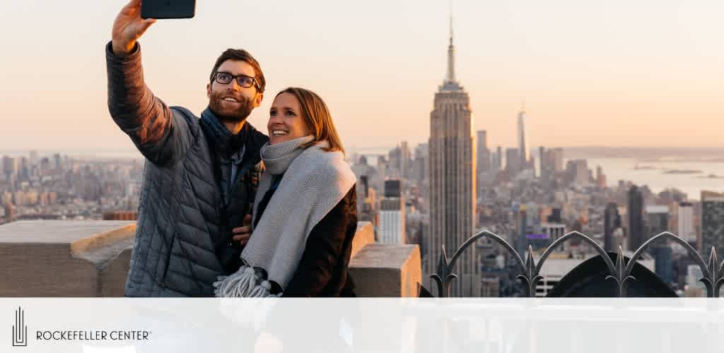 Image shows a man and woman taking a selfie at the Rockefeller Center with the New York City skyline in the background during dusk. The Empire State Building is prominent in the cityscape. They appear joyful, wearing warm clothes. The photo includes the Rockefeller Center's logo.