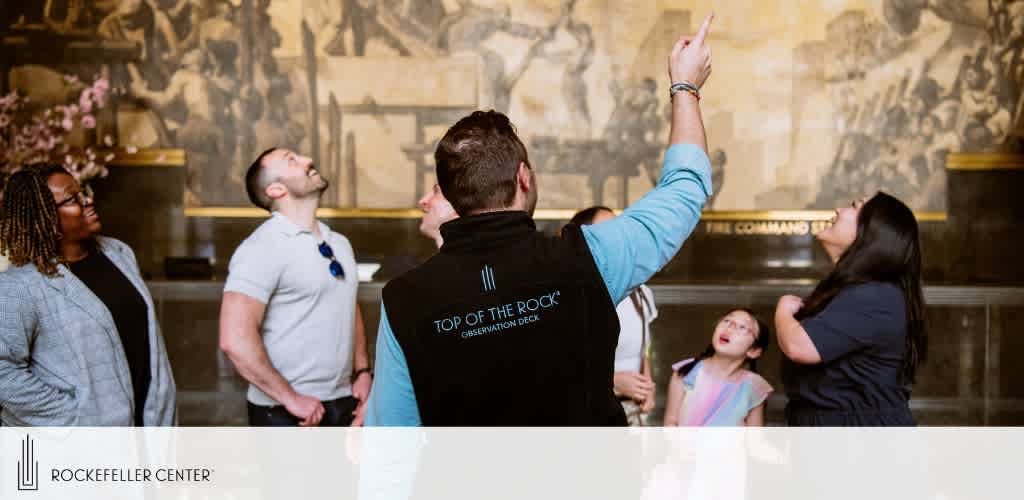 A group of visitors is engaged in a tour at Rockefeller Center. One person, wearing a vest labeled 'Top of the Rock, Observation Deck', is pointing upwards, drawing the group’s attention to details of the mural above. The diverse group looks on with interest and curiosity, capturing the essence of a guided educational experience.