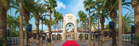 Panoramic view of a theme park entrance with a towering white archway. A red carpet extends toward the gate, flanked by rows of palm trees under a clear, blue sky. Quiet ambiance with no visible visitors.