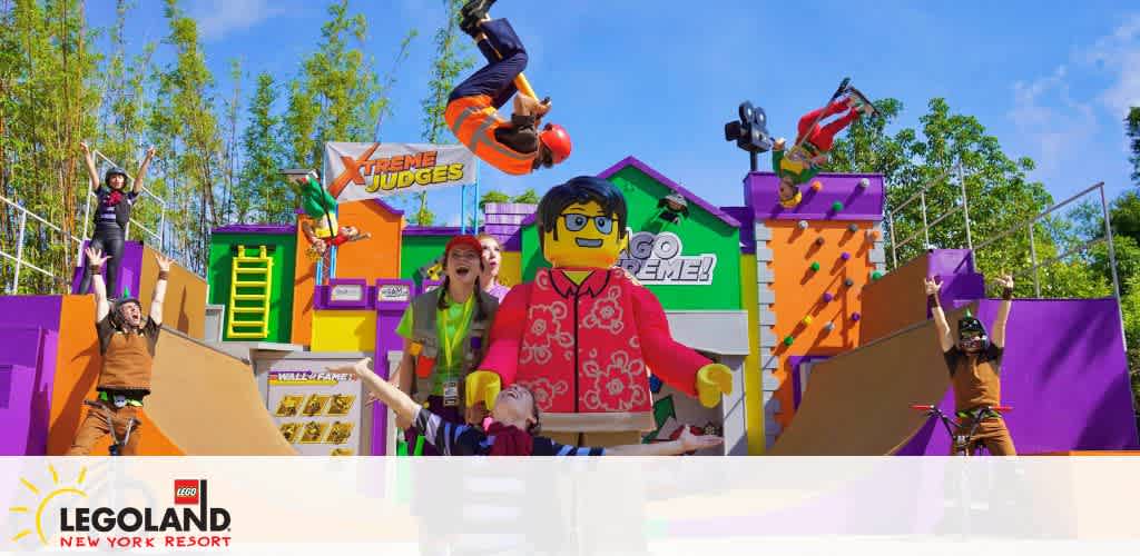 Image displays an energetic scene at LEGOLAND New York Resort with performers and large LEGO figures. A performer is mid-air atop vibrant, colorful ramps while others strike dynamic poses. The backdrop includes oversized LEGO elements and bright decorations under a clear blue sky.