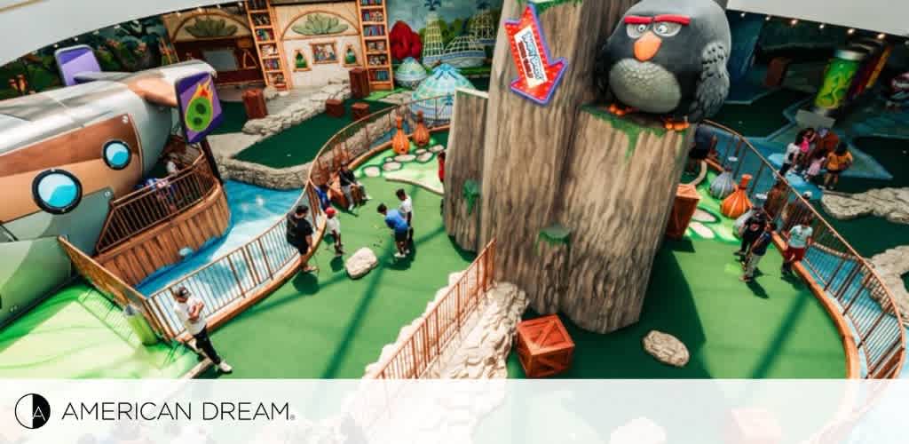 An indoor play area at American Dream with a whimsical theme. To the left, a group of children play near a large, cartoonish airplane. The central area features artificial green flooring, tree stump pathways, and a large sculpted tree with a gray, eagle-like character perched on top. People navigate through the play structures with a background of colorful, oversized bookshelves.