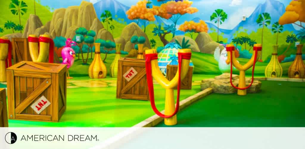 Colorful cartoon-style image showcasing a vibrant mini-golf course with a fantasy theme, including whimsical obstacles like animated trees, oversized TNT crates, slingshots, and colorful vegetation. The American Dream logo is visible in the corner.