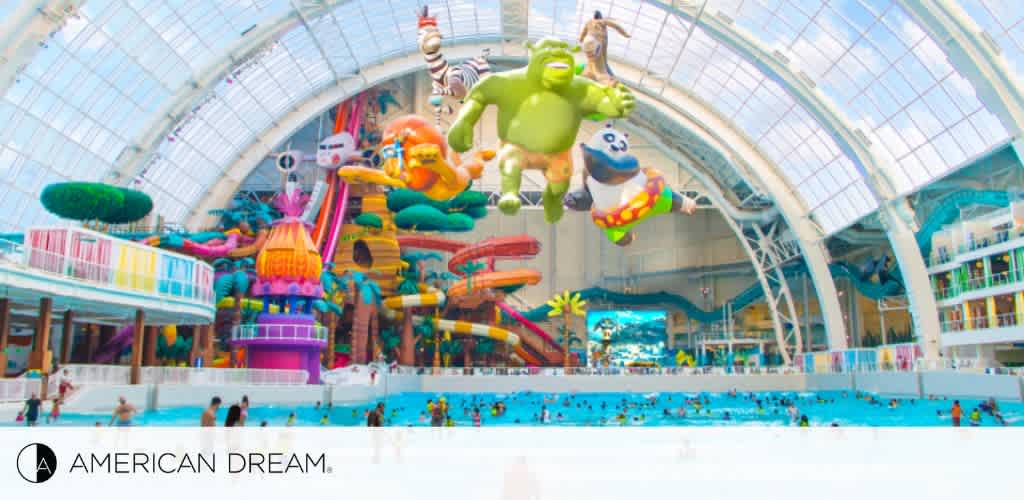 Indoor water park with a lively and colorful atmosphere featuring a large swimming pool and various water slides. Brightly colored, whimsical inflatables are suspended mid-air. A glass dome lets in daylight. The American Dream logo is in the bottom left corner.