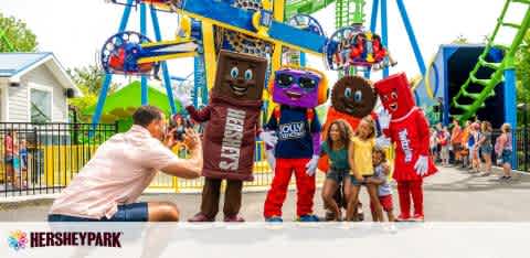 A person kneels to take a photo of a child with four costume characters representing various candy bars at Hersheypark. They stand in front of a rollercoaster, with park visitors in the background. The weather is sunny, and the atmosphere seems festive.