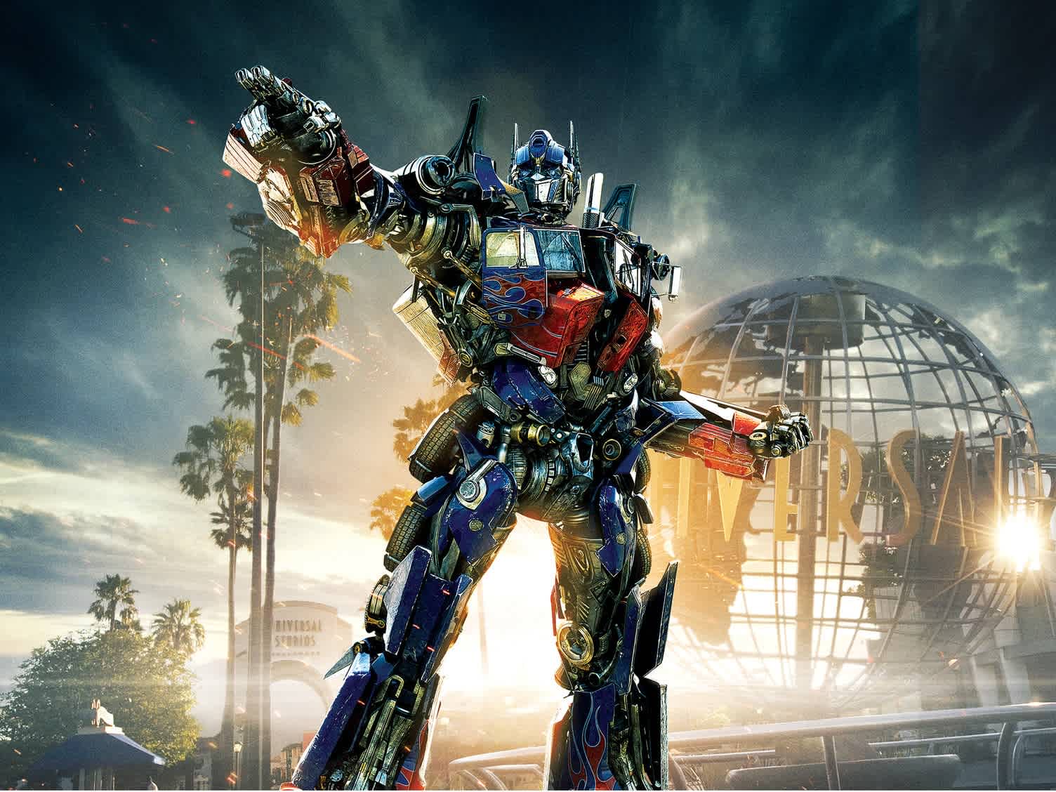 Image shows a large, colorful robot in a heroic stance with one arm raised, set against a sky with clouds. Faint outlines of palm trees are visible in the background suggesting a tropical setting. The robot is intricately detailed with red and blue sections, and there are subtle sparks around it. A spherical structure can be seen on the right.