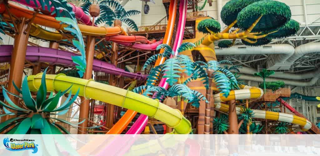 Image shows a vibrant indoor water park with a collection of twisting, multicolored water slides amid artificial palm trees. The slides in yellow, orange, and pink spiral around a structure, creating a playful atmosphere for aquatic fun.