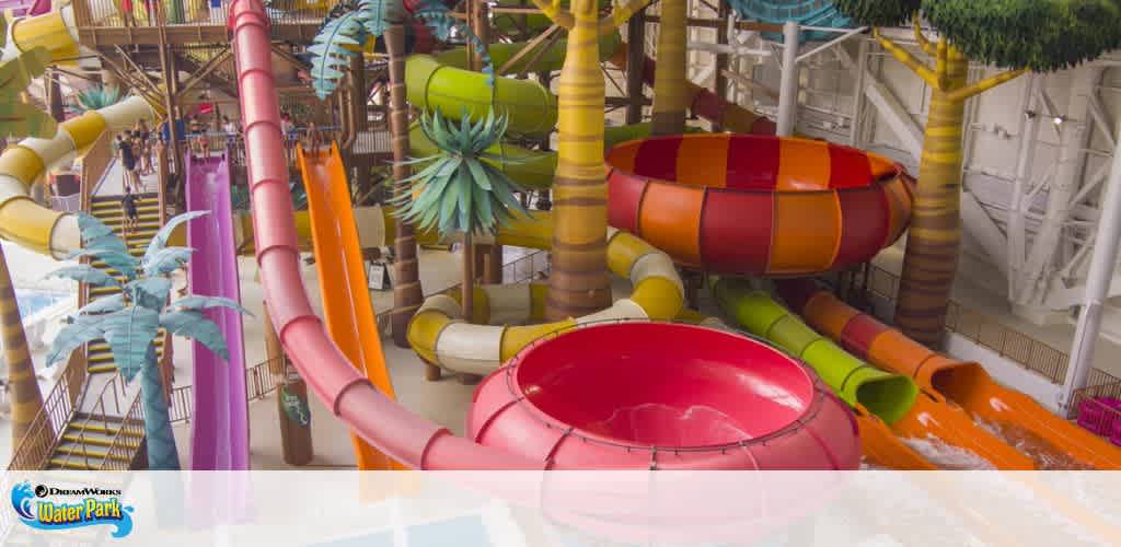 Indoor water park featuring a variety of colorful slides, including a red bowl slide, intertwine amidst palm-tree-like structures. The image showcases a fun and playful atmosphere with slides in orange, yellow, and pink, and a backdrop of supporting beams and a translucent roof allowing natural light.