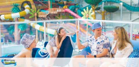 Photo depicts a group of four people relaxing in a water park. Two are sitting facing each other, engaged in conversation, while the other two look on. The background features colorful water slides and palm trees, indicative of a cheerful, leisurely atmosphere typical of a water park setting.