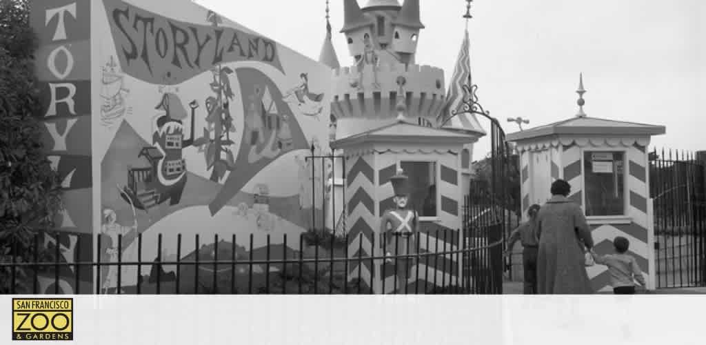 Black and white photo, featuring a mural labeled 'Storyland' with whimsical characters at the San Francisco Zoo & Gardens. A castle with turrets appears to the right and visitors, including a child and adult, are seen near the entrance with a uniformed attendant.