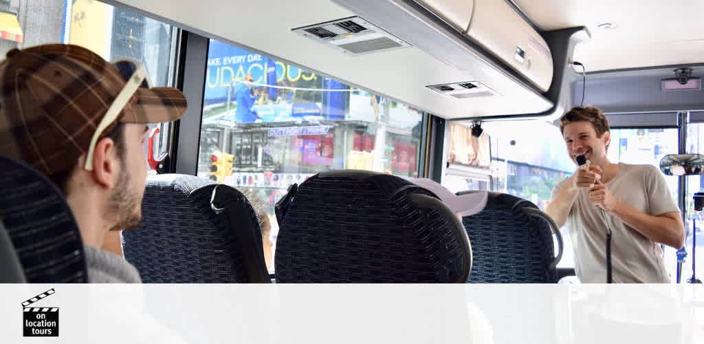 Inside a tour bus, passengers enjoy a guided tour. A man wearing a light-colored shirt and holding a microphone stands in the aisle, smiling as he addresses the group. The interior features rows of dark fabric seats. Daylight streams in through large windows showcasing cityscape advertisements.