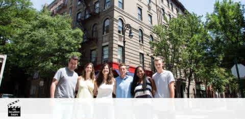 A group of six adults stands smiling behind a white half-wall on a sunny day with trees and a classic five-story brick building in the background. They are casually dressed, with some in light-colored tops and others in darker hues.