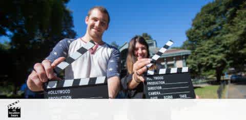 Two people stand outdoors holding film clapboards with a smile. The person on the left is a man in a light shirt with suspenders, and the person on the right is a woman partially obscured by the man, in darker attire. Both are poised as if about to snap the clapper shut. The background is a sunny, tree-lined setting.