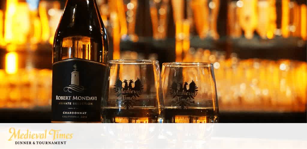 In focus are two wine glasses adorned with the Medieval Times logo, accompanied by a bottle of Robert Mondavi Private Selection Chardonnay positioned behind them. A soft, blurred background evokes a cozy atmosphere. Below, the image is anchored by the Medieval Times Dinner & Tournament logo.