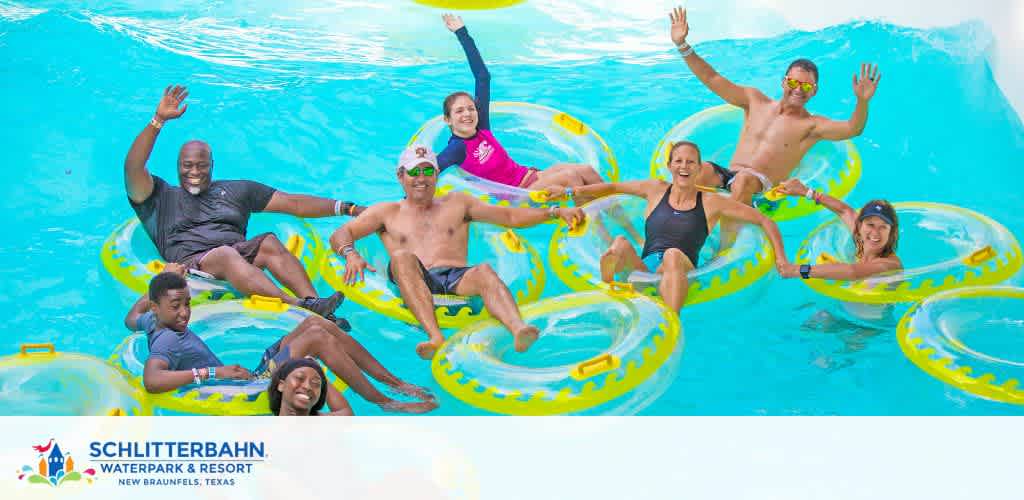 Group of eight happy people of varied ages and ethnicities on yellow inner tubes in a water park slide, hands raised, amidst a splash of aqua-blue water.