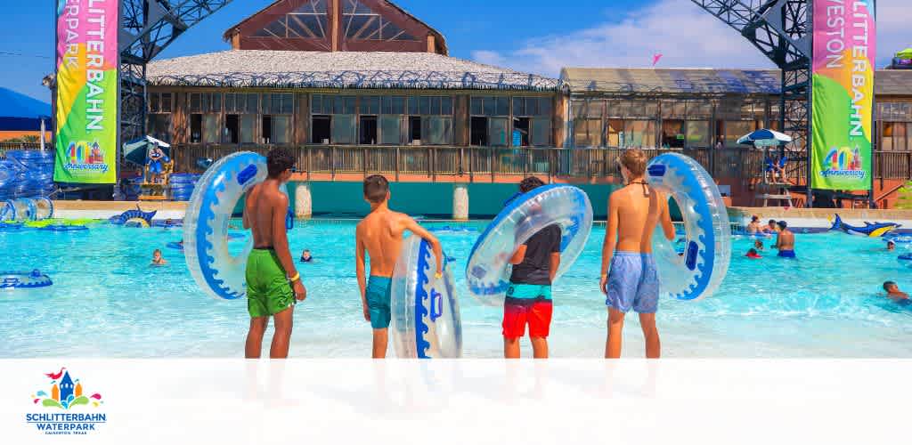 Four young people stand at the edge of the wave pool, each holding a blue inflatable tube, with guests enjoying the water and the water park's main structure in the background on a sunny day.