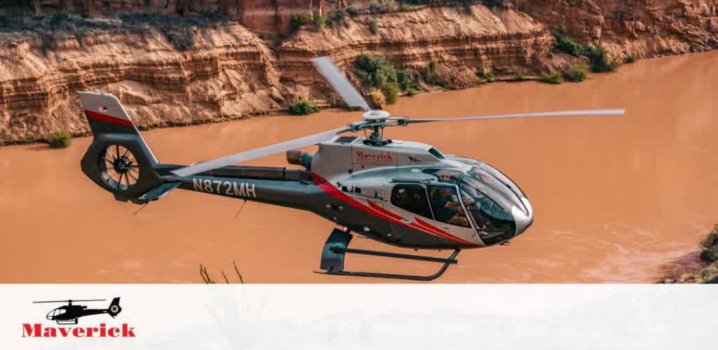 Image of a Maverick helicopter with registration N872MH, in mid-flight over a muddy river, with steep earth-colored cliffs in the background. The helicopter is predominantly white with red and gray accents. The Maverick logo is visible on the side of the chopper and on the tail fin.