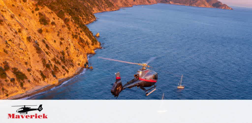 Photo of a red and gray helicopter flying over a coastal area with a steep hillside on the left and open sea on the right. A lone sailboat is visible on the clear blue water. The helicopter is in focus and dominates the foreground while the coastline stretches into the background under a warm, soft light of what appears to be late afternoon. The Maverick logo is visible at the bottom of the image.