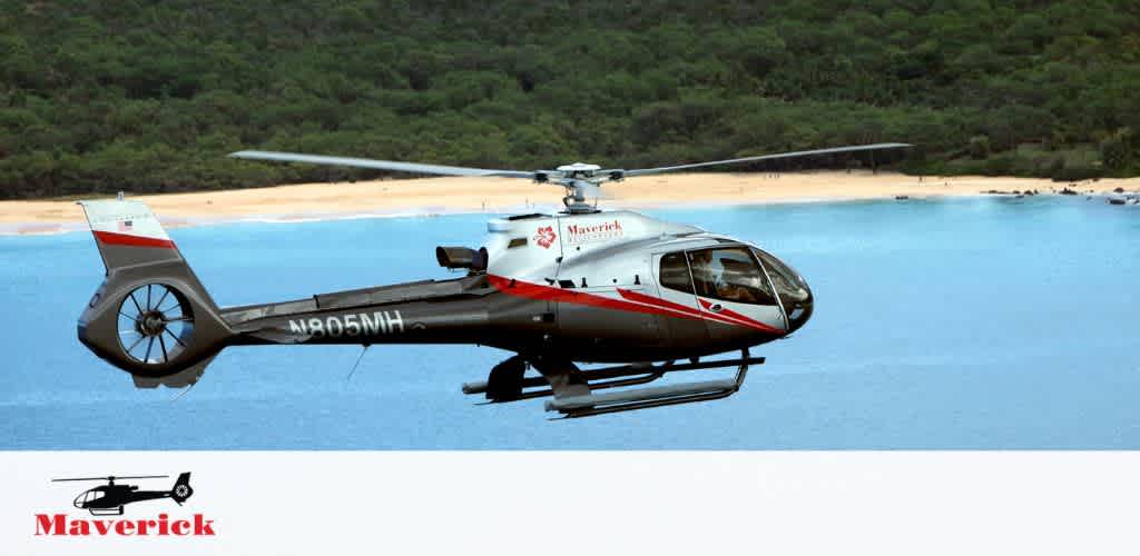A red and white helicopter with the word Maverick on the side hovers above a calm body of water. A lush green hill slopes up in the background, and a sandy beach is visible. The tail of the helicopter displays an identification number. The logo of Maverick with a helicopter silhouette is also shown at the bottom of the image.