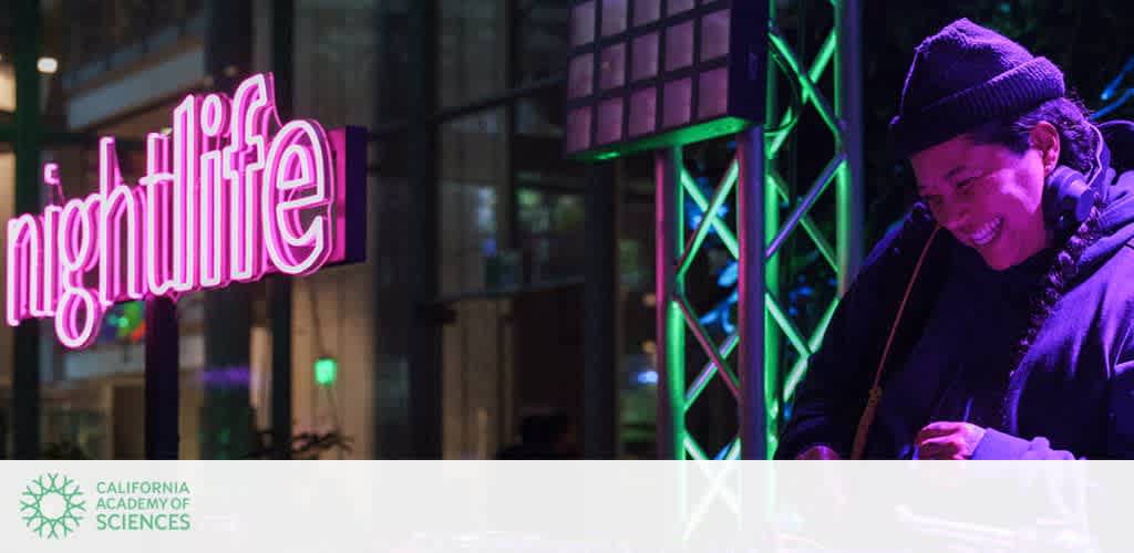 A DJ wearing a beanie is enjoying music under vibrant neon lights at a nighttime event. The word nightlife glows in pink neon, creating an energetic atmosphere. Green and purple lights illuminate the scene, adding to the lively ambiance. The background shows a modern architectural setting, suggesting a fun and engaging environment.