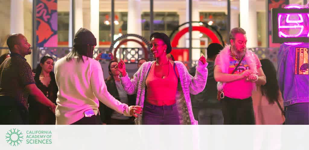 People enjoy dancing at an indoor event, surrounded by colorful lights and vibrant decor. The atmosphere is lively and social, with individuals of various backgrounds smiling and moving to the music. The setting conveys a sense of community and celebration.