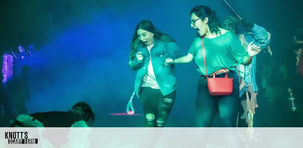Two people react with surprise and laughter in a dimly lit, foggy environment at Knott’s Scary Farm. They are being startled by a character crouched on the ground in front of them, adding to the excitement and thrill of the setting. The lighting is vivid with green and blue hues.