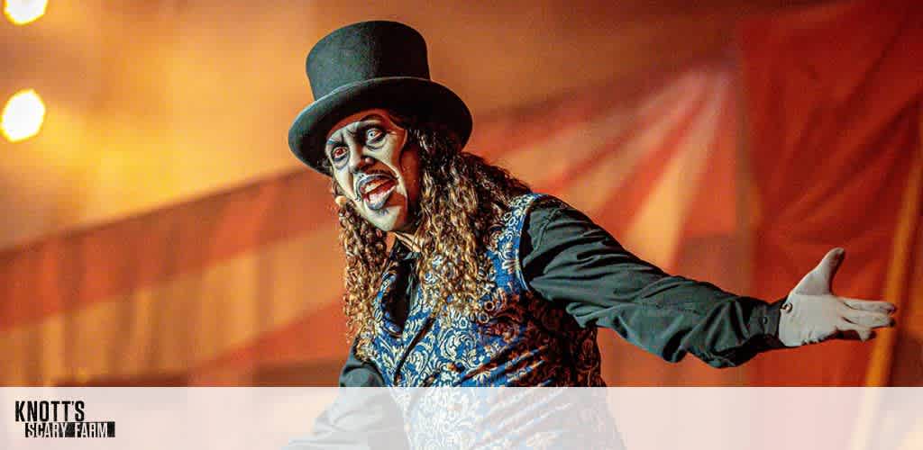 A performer dressed in a top hat, black shirt, and ornate vest gestures dramatically on stage with a painted face. The background features warm lighting and blurred stripes, suggesting a carnival or circus theme. The atmosphere is theatrical, with an eerie twist, as part of a spooky entertainment event.