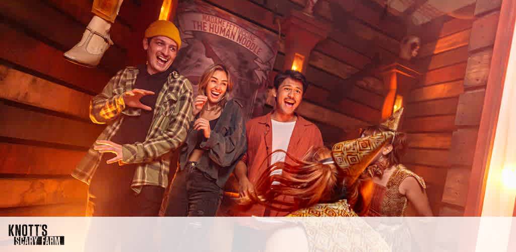 A group of four people are inside a dimly lit, haunted-themed room at Knott's Scary Farm. They appear surprised and amused, reacting to a spooky scene. The room is decorated with wooden panels and creepy figures. A poster for a magician called The Human Noodle is visible in the background, adding to the eerie ambiance.