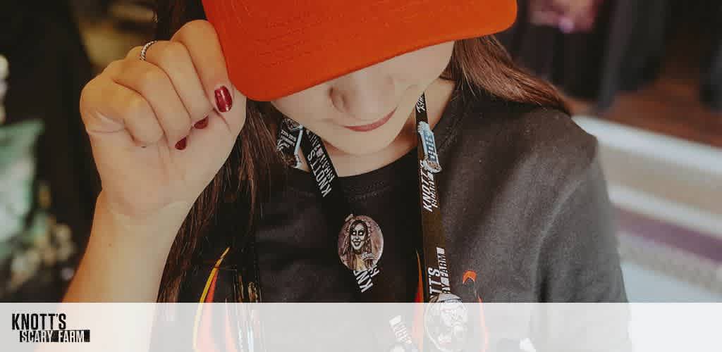 A person wearing a red cap slightly tilted downwards, with a black lanyard featuring a Knotts Scary Farm badge. The person has long brown hair and is dressed in a black shirt. The setting appears to be indoors with blurred background elements. The image showcases a casual, themed attire associated with Knotts Scary Farm.