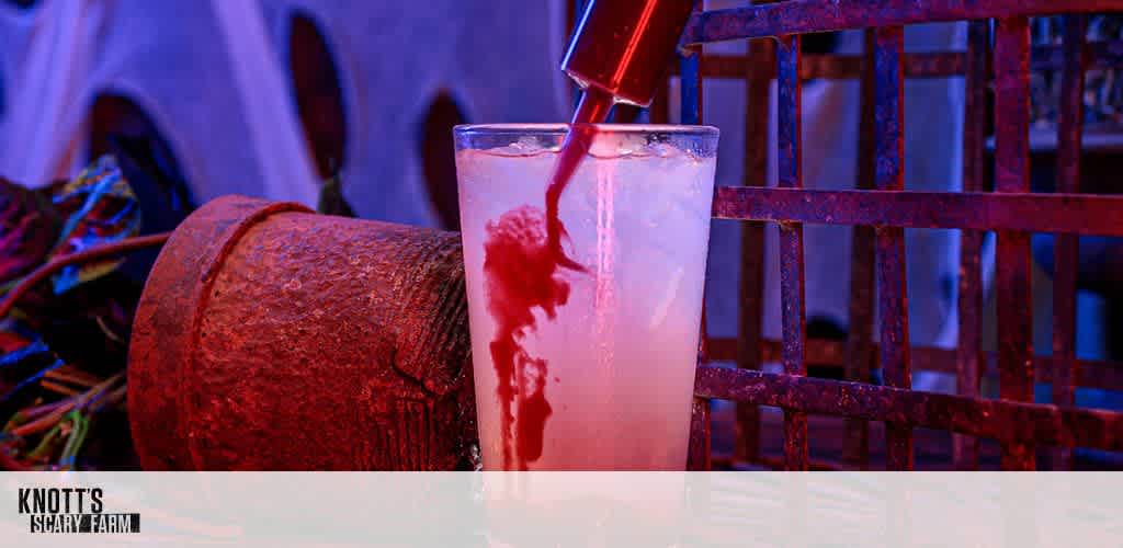 A spooky drink being prepared at Knott's Scary Farm. A tall glass is being filled with a red liquid from a spout, creating a dramatic contrast with the light-colored liquid already in the glass. In the background, eerie decor adds to the Halloween theme, featuring a rustic metal grid and scattered props, enhancing the mysterious atmosphere of the scene.