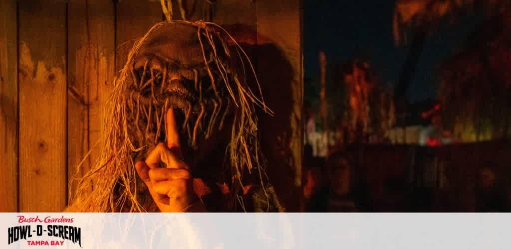 Person wearing a scarecrow mask with a finger to lips sign, standing against a wooden background at night during Halloween event