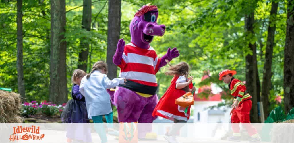 Image shows a playful outdoor scene with a person in a large purple dragon costume interacting with children. The costume character waves hello, as colorful flowers and lush trees create a vibrant background. One child wears a red firefighter outfit, adding to the lively and whimsical atmosphere. The words 'Idlewild HALLOWBOO!' are visible in the corner, indicating a festive event.