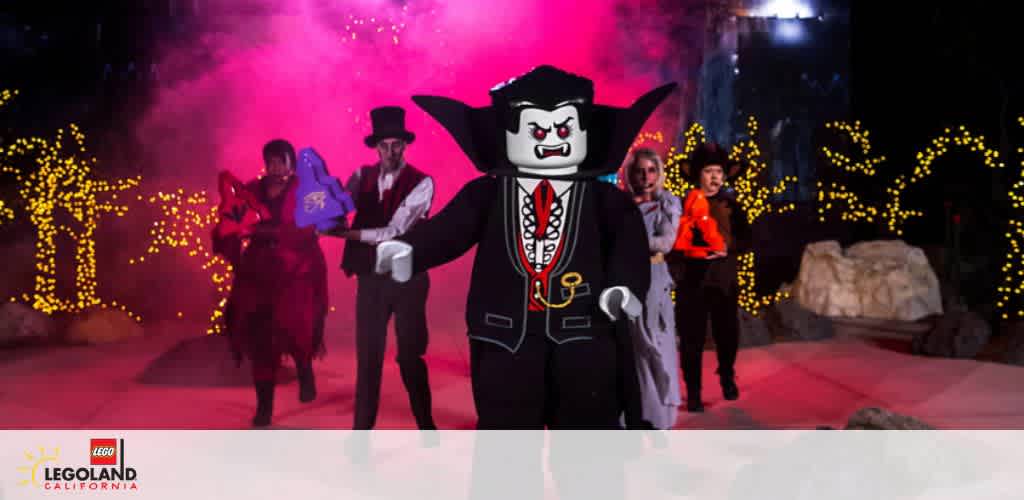 Image features a lively stage scene with a large figure in a vampire costume at the forefront and entertainers in background. They stand before a backdrop of twinkling lights, conveying a dynamic, festive atmosphere. The LEGOLAND California logo is visible, suggesting the event's location.