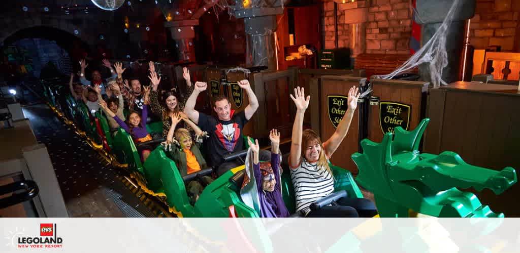 People on a green dragon-themed roller coaster in a dark, castle-like indoor ride at Legoland New York Resort, with hands raised in excitement.