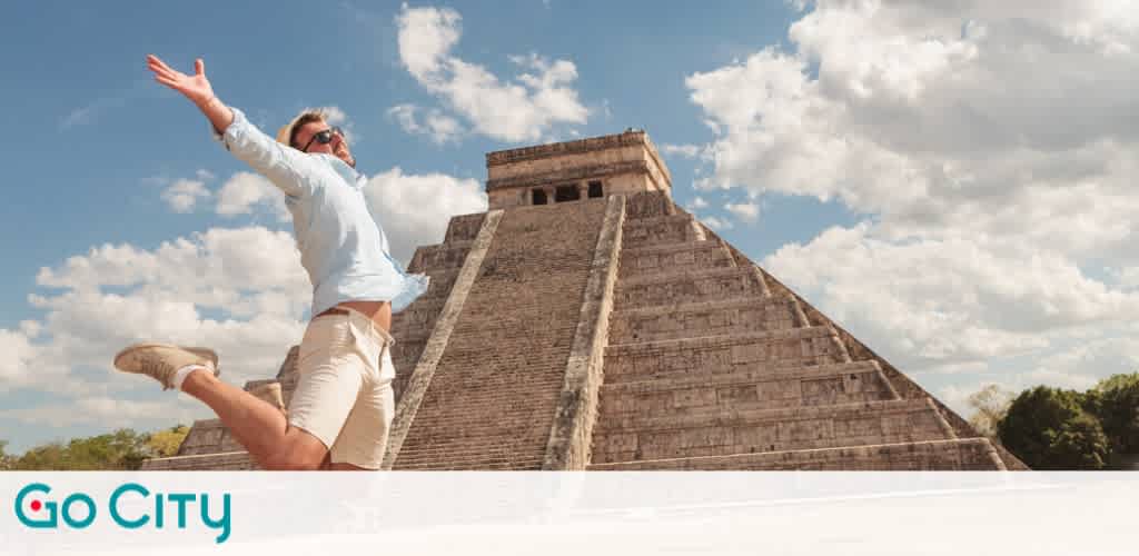 A person jumps joyfully in front of the ancient stepped pyramid of Chichen Itza under a partly cloudy blue sky. The person wears casual light clothing and sunglasses, expressing excitement and freedom. The historic stone structure stands tall in the background, showcasing its intricate architecture. A logo for Go City is visible in the bottom corner of the image.