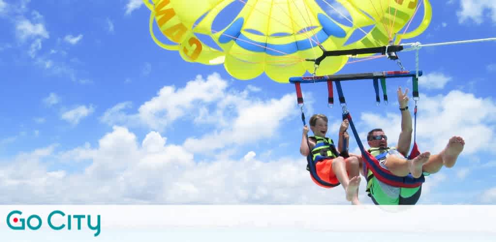 Two people enjoying parasailing under a bright yellow parachute against a clear blue sky. They are harnessed securely, experiencing an adventurous ride high above the sea. The ocean is visible below them, adding to the thrilling and scenic experience. Perfect for an exciting outdoor adventure.
