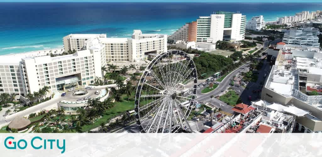 Aerial view of a coastal city featuring a large Ferris wheel in the center. Surrounding the Ferris wheel are several modern multi-story hotels and buildings. The ocean spans the top of the image with its vibrant blue waters, contrasting with the white sandy beach. A curved road with light traffic winds through the landscape, bordered by lush greenery. The image includes the Go City logo at the bottom left corner.