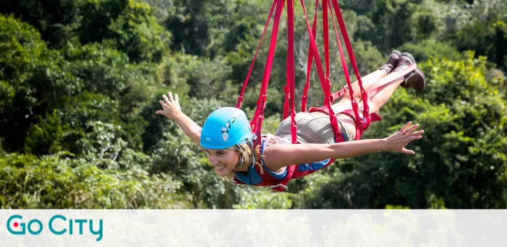A person is enjoying an outdoor zip-lining adventure wearing a blue helmet and secured in a red harness. They are flying in a horizontal position with arms outstretched, set against a lush green forest backdrop. The scene is vibrant and energetic, showcasing the thrill of the experience. Go City logo is at the bottom left corner.