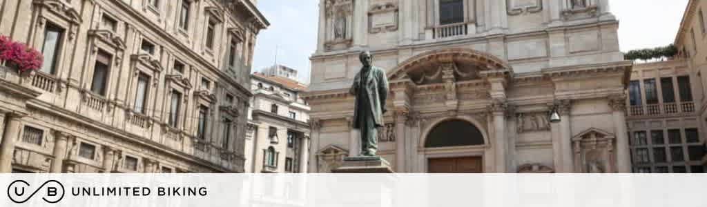 A historical European plaza with a prominent statue in the center, surrounded by ornate classical buildings. The architecture features columns and detailed facades, suggesting an old-world charm. The logo and text Unlimited Biking is at the bottom left corner, indicating a biking service or company associated with the scene.