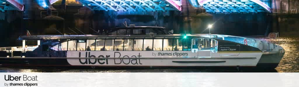 A sleek, modern ferry labeled Uber Boat by Thames Clippers is docked at night. Soft light reflects on the water, highlighting the boat's glass windows and sleek white exterior. Above, a brightly lit bridge with blue tones creates a dramatic backdrop.
