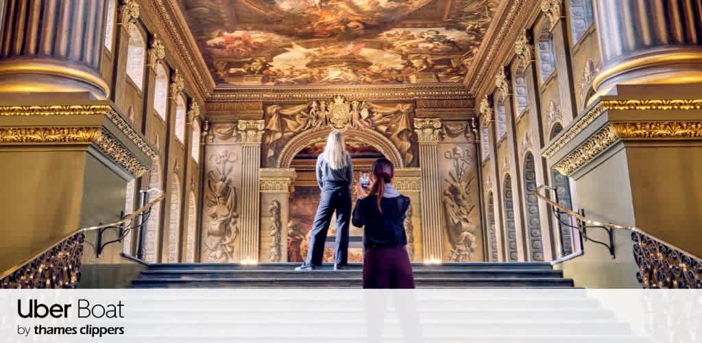 Two people stand on ornate steps in a grand hall, admiring a lavishly painted ceiling depicting historical scenes. The hall features tall, decorated columns and intricate wall carvings, evoking a sense of classic elegance and artistic grandeur. Sunlight streams in through tall windows, enhancing the rich colors of the artwork.