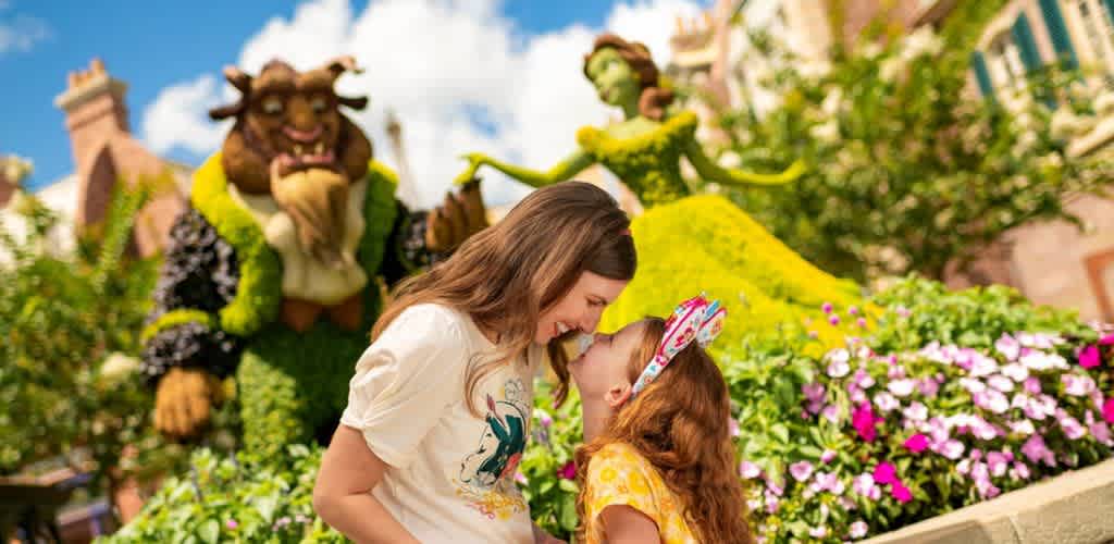A woman and a young girl share a joyful moment in front of large topiary sculptures resembling a beast and a woman in a vibrant garden setting. The woman leans down to touch noses with the girl, who wears a playful headband. Bright flowers and greenery surround them, creating a whimsical and colorful scene. The sculptures in the background add a sense of enchantment to the sunny day.