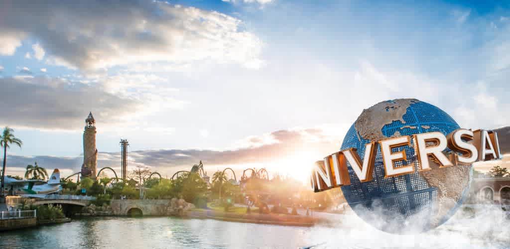Sunny landscape featuring a large globe statue with the word Universal across it. The background includes a theme park with roller coasters, tall structures, and lush greenery, beside a calm water body reflecting the park and clear sky. The scene captures a vibrant, adventurous atmosphere with sunlight casting a warm glow over the entire area, perfect for a day of fun and excitement at the park.
