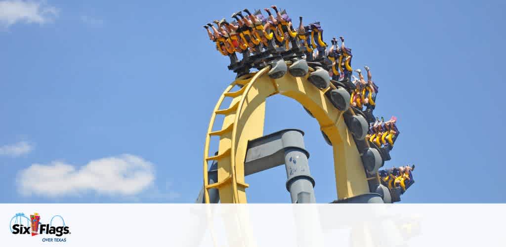 A thrilling roller coaster with bright yellow tracks at Six Flags Over Texas twists upside down against a blue sky. Riders are seen with arms raised, experiencing an exhilarating upside-down loop. The image captures the excitement and energy of the amusement park ride. At the bottom left, the Six Flags Over Texas logo is visible.