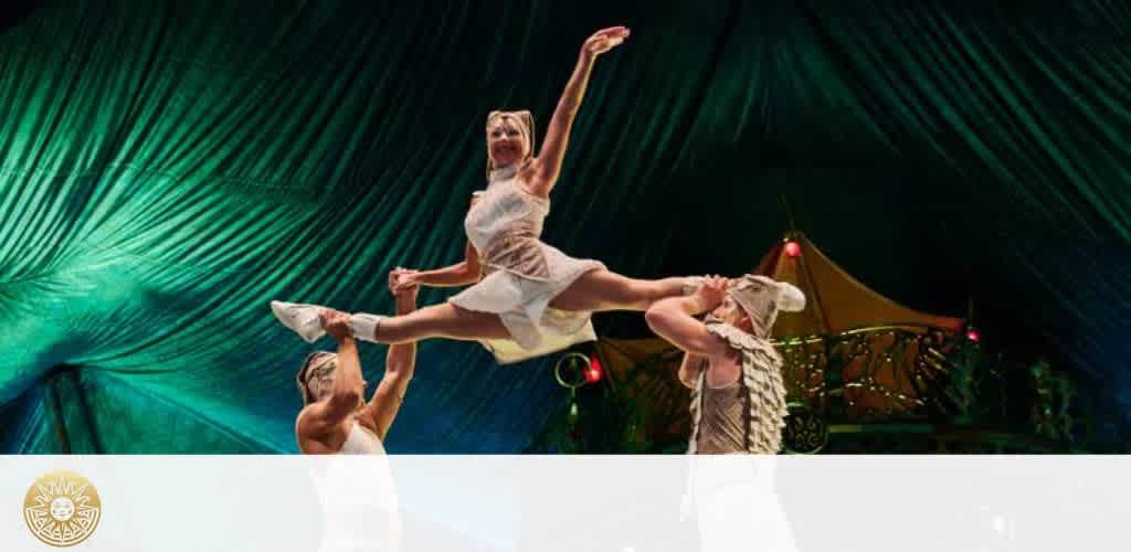 Three performers in a vibrant circus scene. Two individuals on the ground support a third performer, who is elegantly leaping in the air, wearing a light outfit. The background features a dramatic, teal-colored draped ceiling that adds to the dynamic atmosphere. Warm lighting enhances the image, creating a sense of excitement and spectacle.