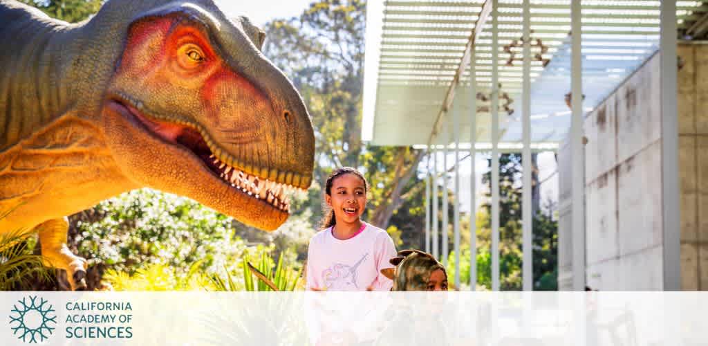 A smiling child stands next to a large dinosaur model in a sunny outdoor area with greenery. The scene takes place under a modern canopy at a science museum. The setting is lively and educational, inviting visitors to explore and learn.