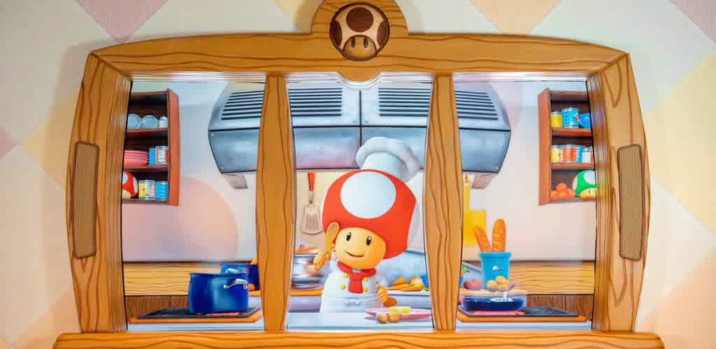 A cheerful cartoon character with a red and white mushroom hat stands behind a wooden framed kitchen window. The kitchen is equipped with blue pots, cooking utensils, and colorful shelves holding ingredients. The scene is playful and vibrant, with bright colors and a whimsical design, creating a fun and inviting atmosphere.