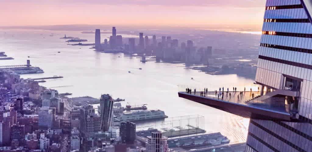 A stunning view of a city skyline at sunset is seen from a unique vantage point. The image shows a tall building with a glass observation deck extending outward, where people are standing and enjoying the expansive vista. The foreground reveals a sprawling urban landscape with diverse architecture, while the background features a body of water and another cityscape under a warm, colorful sky.