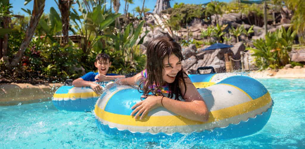 Children enjoy a sunny day at a water park, floating on colorful inflatable tubes in a lazy river. Surrounded by lush greenery and tropical plants, they smile and splash as they drift through the bright blue water. The backdrop features rocky landscapes and clear skies, creating a fun and relaxing atmosphere perfect for family-friendly recreation.