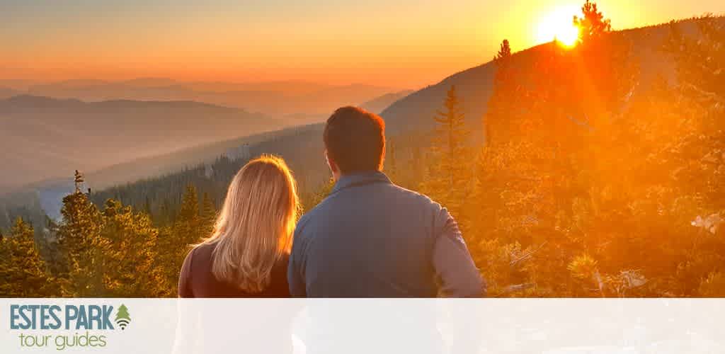 A couple stands on an overlook, gazing at a stunning sunset over a forested mountain range. The sky is painted in warm hues of orange and gold, casting a serene glow on the landscape. The scene exudes tranquility and natural beauty, inviting viewers to experience the breathtaking vistas of Estes Park.
